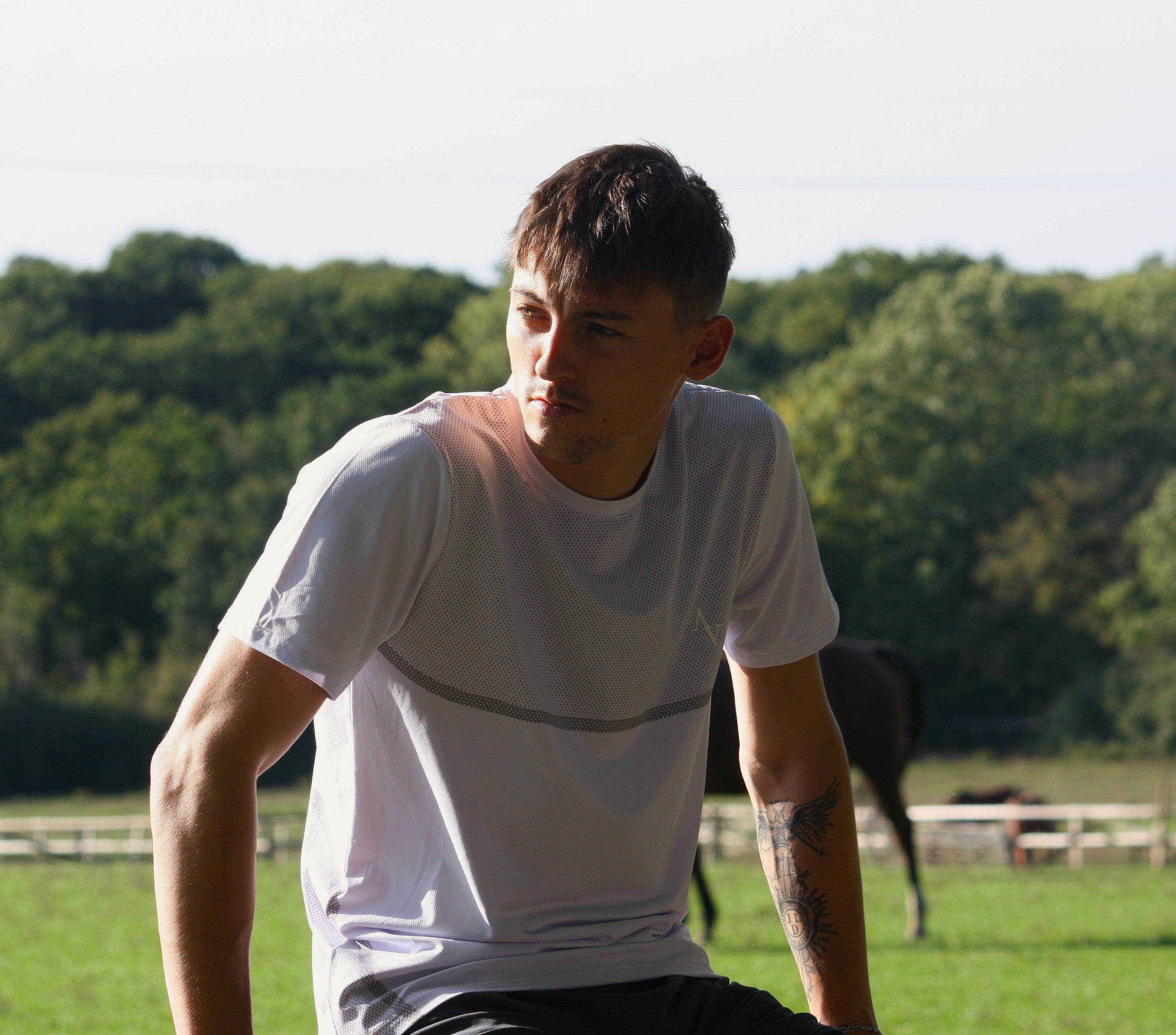Man in a white t-shirt sitting on a grassy field with trees in the background